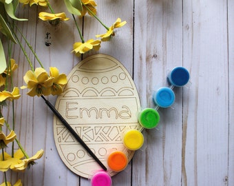 May include: Wooden Easter egg shaped craft with the name "Emma" written on it. The egg is surrounded by six small paint pots in rainbow colors, including red, orange, yellow, green, blue, and purple.