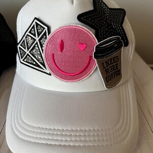 May include: White mesh trucker hat with a pink smiley face, a diamond, a black star, and a brown coffee cup patch that says "I need more coffee".