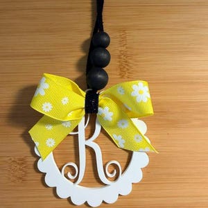 Pu&ograve; includere: Un ornamento con monogramma bianco con bordo smerlato con la lettera "R" &egrave; accentuato da un nastro giallo con motivi a margherita bianca. L'ornamento &egrave; appeso a un cordoncino nero con tre perline di legno nere.