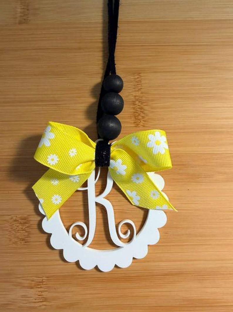 Pu&ograve; includere: Un ornamento con monogramma bianco con bordo smerlato con la lettera "R" &egrave; accentuato da un nastro giallo con motivi a margherita bianca. L'ornamento &egrave; appeso a un cordoncino nero con tre perline di legno nere.