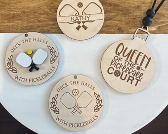 Può includere: Tre ornamenti in legno con tema pickleball. Un ornamento è rotondo e dice "Queen of the Pickleball Court". Un altro ornamento è rotondo e dice "Deck the Halls with Pickleballs". Il terzo ornamento è rotondo e dice "Kathy" con un design di racchetta da pickleball.
