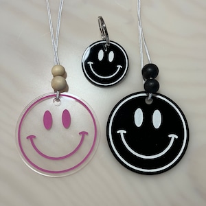 May include: Two clear acrylic smiley face necklaces with pink outlines and black beads. One necklace has wooden beads. The other necklace has a black smiley face keychain.