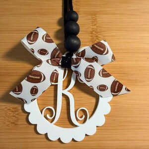 Pu&ograve; includere: Un ornamento bianco con l'iniziale monogramma "K" con un bordo smerlato, adornato con un fiocco con nastro a tema football. L'ornamento &egrave; sospeso da un cordoncino nero con tre perline nere.