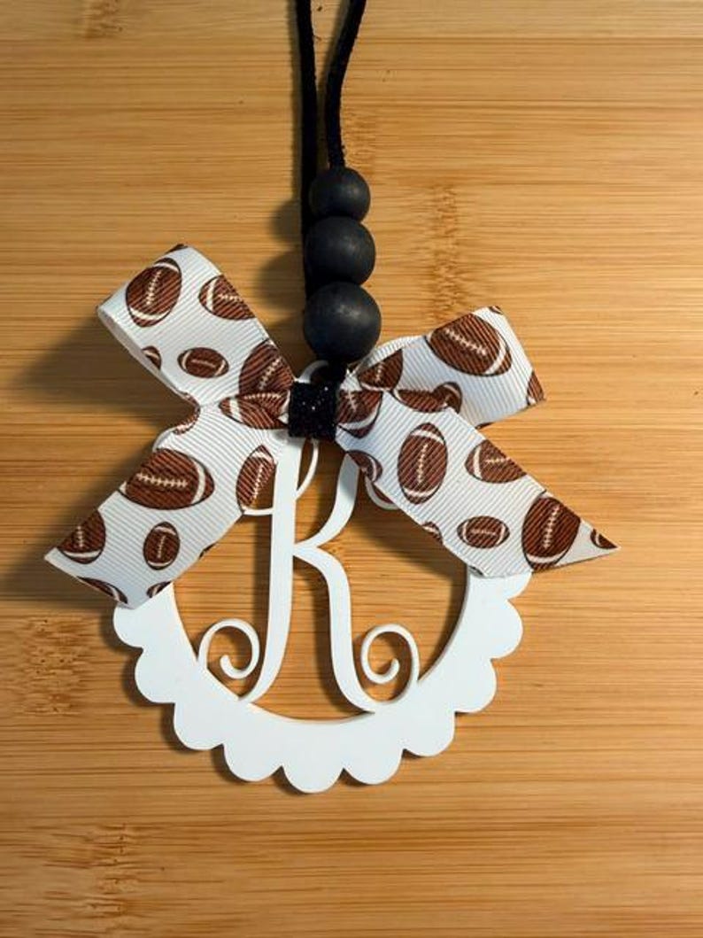 Pu&ograve; includere: Un ornamento bianco con l'iniziale monogramma "K" con un bordo smerlato, adornato con un fiocco con nastro a tema football. L'ornamento &egrave; sospeso da un cordoncino nero con tre perline nere.