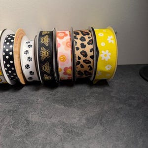 Pu&ograve; includere: Una collezione di rocchetti di nastro decorativo in vari modelli e colori. I disegni includono pois, impronte di zampe, api, fiori e stampa leopardata. I nastri sono nelle tonalit&agrave; di nero, bianco, rosa, giallo e marrone.
