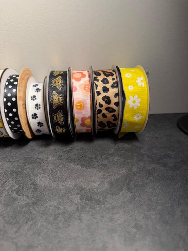 Pu&ograve; includere: Una collezione di rocchetti di nastro decorativo in vari modelli e colori. I disegni includono pois, impronte di zampe, api, fiori e stampa leopardata. I nastri sono nelle tonalit&agrave; di nero, bianco, rosa, giallo e marrone.