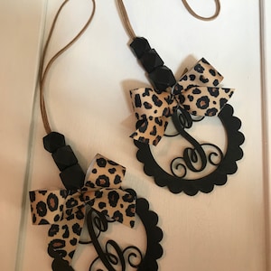 Pu&ograve; includere: Due ciondoli per auto con monogramma nero con bordo smerlato, fiocco con stampa leopardata e cordino marrone. I ciondoli hanno una lettera "C" in corsivo. I ciondoli sono appesi su uno sfondo bianco.