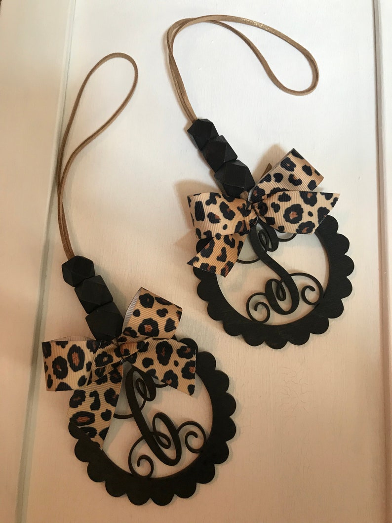 Pu&ograve; includere: Due ciondoli per auto con monogramma nero con bordo smerlato, fiocco con stampa leopardata e cordino marrone. I ciondoli hanno una lettera "C" in corsivo. I ciondoli sono appesi su uno sfondo bianco.