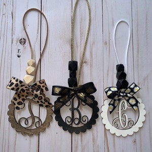 May include: Three wooden monogram ornaments with scalloped edges and decorative bows. One has a leopard print bow, another a black bow with gold accents, and the third a white bow with daisy accents. Each has a different letter.