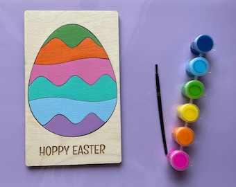 May include: Wooden Easter egg shaped craft with a multi-colored design and the text "Hoppy Easter". The craft comes with a set of seven paint pots in various colors and a paintbrush.