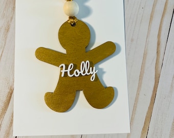 Può includere: Ciondolo a forma di uomo di pan di zenzero dorato con il nome "Holly" scritto su di esso. Il ciondolo è appeso a due perline di legno bianche.