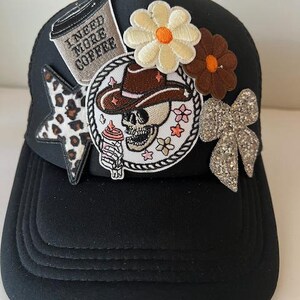 May include: Black trucker hat with decorative patches. Patches include a coffee cup with "I NEED MORE COFFEE", a star with a leopard print, a skull wearing a cowboy hat, flowers, and a glitter bow. The hat has a curved brim.