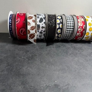 Pu&ograve; includere: Una collezione di rocchetti di nastro decorativo con vari motivi. I disegni includono stelle, paisley, palloni da football, stampa mucca, glitter, margherite, quadretti, palline da baseball e da tennis. I nastri sono su rocchetti bianchi.