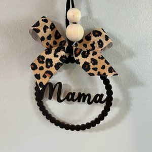 May include: A black wooden circle with the word "Mama" cut out, decorated with a leopard print bow and wooden beads hanging from a black cord.