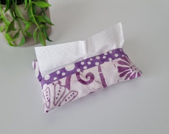 May include: A patterned fabric tissue holder in purple and white, with white tissues protruding from the top. The holder is rectangular and made of fabric with a floral design. A small green plant is visible in the background.