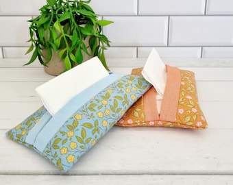 May include: Two fabric tissue box covers. One is light blue with yellow floral pattern, the other is orange with pink floral pattern. Both have a rectangular shape with a slit for tissue dispensing. A green plant in a pot is in the background.