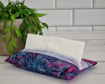 May include: A patterned fabric tissue holder in blue, purple, and pink floral design. White tissues are partially visible, emerging from the top opening. The holder is rectangular and rests on a white surface.