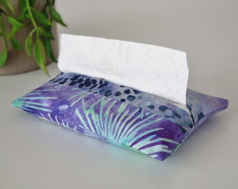 May include: A rectangular tissue box cover in batik fabric, featuring purple, blue, and teal hues. White tissues are partially visible. The fabric design includes a botanical pattern, adding a decorative touch to the tissue box.