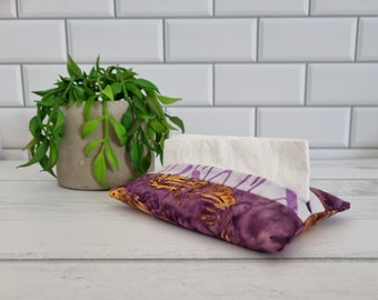 May include: A fabric tissue box cover with a purple and gold batik design. The rectangular cover holds a stack of white tissues. A small potted plant is visible in the background. The cover is designed to fit standard tissue boxes.