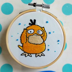 May include: Embroidered hoop art featuring a yellow Pokemon character with brown eyes and a brown nose. The character is surrounded by blue beads on a white background.