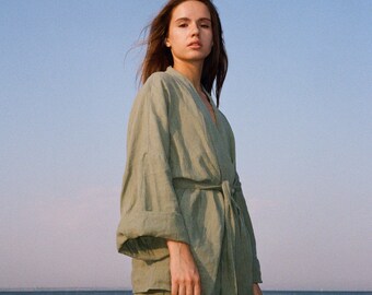 May include: A woman wearing a sage green linen kimono-style top with a matching belt and pants. The outfit is lightweight and flowing, perfect for warm weather. The background features a clear blue sky and the ocean.