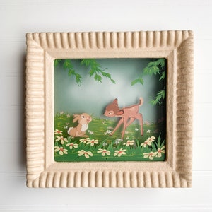 May include: A framed picture with a 3D effect of a fawn and a rabbit in a forest setting. The fawn is brown with white spots and is looking at the rabbit. The rabbit is brown and white and is looking at the fawn. The background is a green forest with flowers.