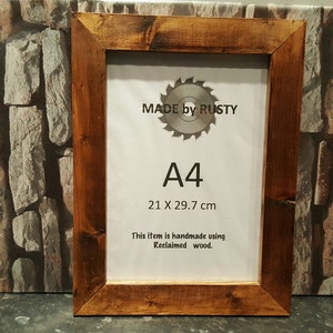 Peut inclure: Cadre photo en bois A4 de couleur marron fonc&eacute; avec une finition rustique. Le cadre est fabriqu&eacute; &agrave; partir de bois de r&eacute;cup&eacute;ration et porte l'inscription "MADE by RUSTY" et "A4 21 x 29,7 cm". Le texte "This item is handmade using Reclaimed wood" est &eacute;galement visible.