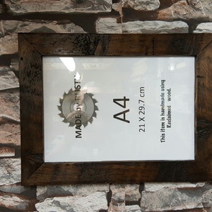 Peut inclure: Un cadre photo rectangulaire en bois brun fonc&eacute;. Il contient une feuille de papier blanche avec le texte "A4 21 x 29,7 cm" et "MADE BY RUST". Le cadre est fait &agrave; la main &agrave; partir de bois de r&eacute;cup&eacute;ration.