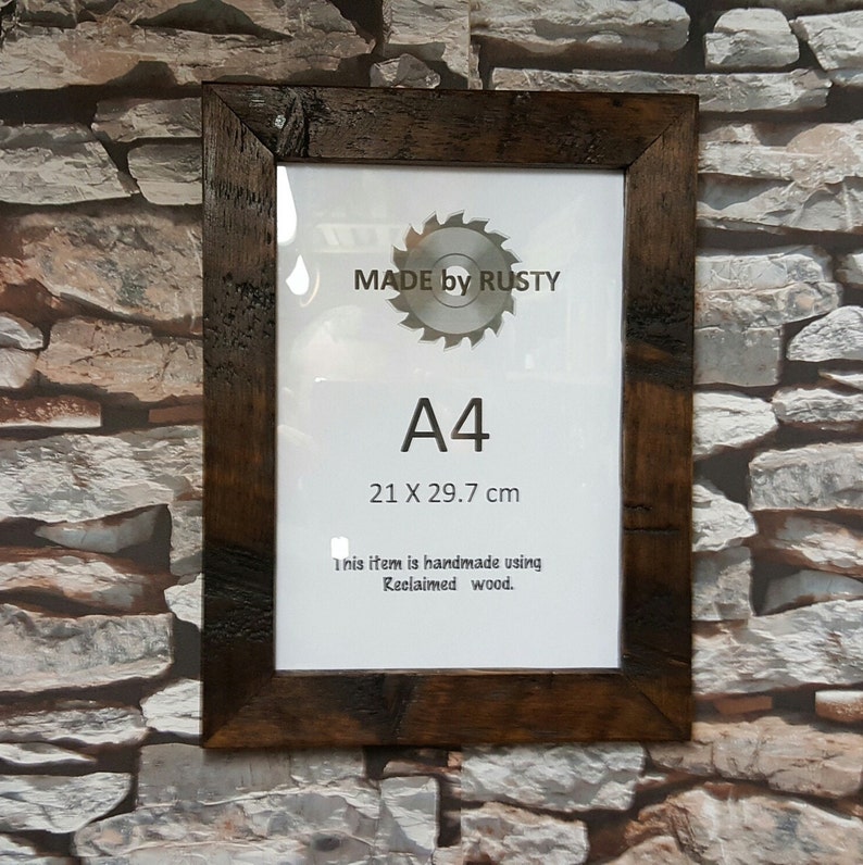 Peut inclure: Cadre photo de format A4 avec un cadre en bois de r&eacute;cup&eacute;ration brun fonc&eacute;. Le cadre contient un papier blanc avec le texte "MADE by RUSTY", "A4" et des dimensions de 21 x 29,7 cm. Le texte "This item is handmade using Reclaimed wood" est &eacute;galement visible.