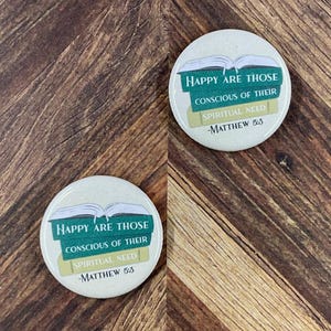 May include: Two round, off-white buttons with the text "HAPPY ARE THOSE CONSCIOUS OF THEIR SPIRITUAL NEED - MATTHEW 5:3" in green and yellow. The buttons feature an open book graphic and are set against a wooden background.