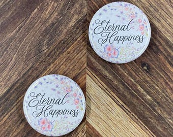 May include: Two round buttons with floral designs and the words "Eternal Happiness" in elegant script. The buttons feature a white background with pink and purple flowers and green leaves. The buttons are on a wooden surface.