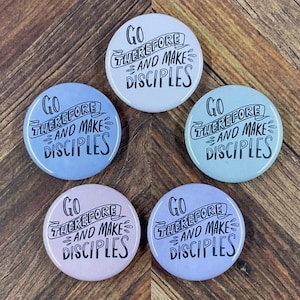 Könnte beinhalten: Fünf runde Knöpfe mit dem Text "Go therefore and make disciples" in schwarzer Schrift auf einem weißen Banner. Die Knöpfe sind in verschiedenen Blautönen und Lila.