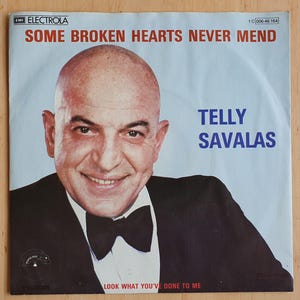 Op de afbeelding: Een vintage vinylhoes met Telly Savalas. De hoes heeft een lichtblauwe achtergrond met de tekst "SOME BROKEN HEARTS NEVER MEND" in het rood. Savalas draagt een zwart pak en een vlinderdas en glimlacht. De tekst "LOOK WHAT YOU'VE DONE TO ME" staat onderaan.