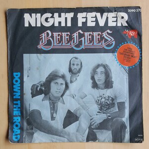 Puede incluir: Una portada de disco de vinilo vintage con una foto de los miembros de la banda Bee Gees. La portada es negra con texto blanco que dice "Night Fever" y "Down the Road". Los miembros de la banda visten ropa informal y sonríen. El disco es de la banda sonora de la película "Saturday Night Fever".