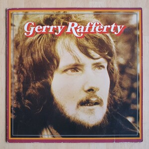 Op de afbeelding: Een vintage platenhoes met een close-up portret van Gerry Rafferty, een Schotse zanger-songwriter. De hoes is warm bruin van kleur met een gele rand en de naam van de artiest in rode letters.