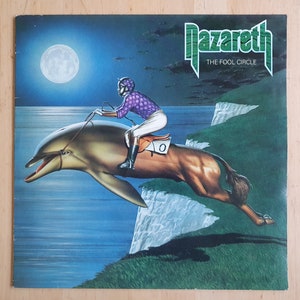 Pode incluir: Uma capa de álbum de vinil vintage com uma pessoa montando um golfinho sobre um penhasco. A capa do álbum é para a banda Nazareth e o título do álbum é "The Fool Circle".