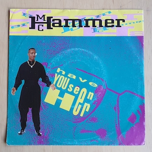 Op de afbeelding: Een blauwe en paarse platenhoes met een gele 'H' en de tekst 'MC Hammer' en 'Have You Seen Her' erop. Een man in een zwart shirt en zwarte broek staat voor de platenhoes.