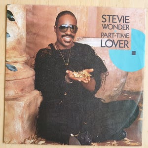 Pode incluir: Capa do álbum de Stevie Wonder com uma foto em preto e branco de Stevie Wonder sentado em um jardim, usando óculos de sol e um suéter preto. O título do álbum "Part-Time Lover" está impresso em branco sobre um fundo azul.