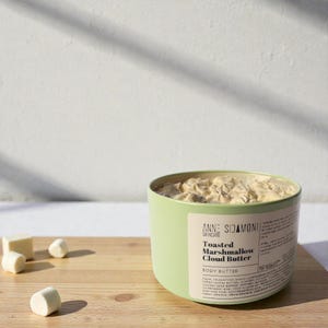 Peut inclure: Un pot vert clair de beurre corporel Anne SIdamoni Skincare Toasted Marshmallow Cloud Butter. Le pot est rempli d'un beurre corporel crémeux brun clair. Plusieurs guimauves blanches et beiges sont éparpillées sur une surface en bois.