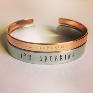 May include: A double-layered bracelet with the words "I'm Speaking" engraved on both bands. The top band is copper and the bottom band is silver.