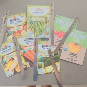 Puede incluir: Una colecci&oacute;n de paquetes de semillas y marcadores de plantas de metal con los nombres de diferentes verduras escritos en ellos, incluyendo pimientos, calabac&iacute;n, zanahorias, frijoles, tomates, pepinos y frijoles de jard&iacute;n. Los paquetes de semillas son todos org&aacute;nicos y presentan una variedad de colores, incluyendo verde, rojo, amarillo y naranja.