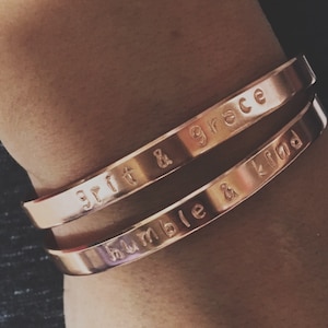 May include: Two gold-toned metal bracelets with engraved messages. The top bracelet reads "grit & grace" and the bottom bracelet reads "humble & kind".