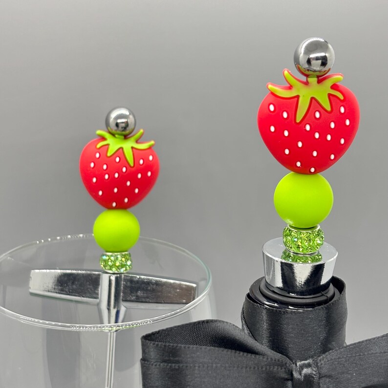 K&ouml;nnte beinhalten: Zwei dekorative Flaschenverschl&uuml;sse mit Erdbeermotiv. Jeder Verschluss hat eine rote Erdbeere mit wei&szlig;en Samen, einen gr&uuml;nen Stiel, eine limettengr&uuml;ne Perle und eine silberne Kugel. Ein Verschluss befindet sich auf einem Weinglas, der andere auf einer Weinflasche.