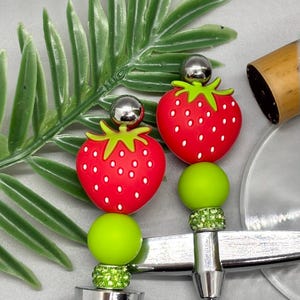 K&ouml;nnte beinhalten: Nahaufnahme von zwei Flaschenverschl&uuml;ssen im Erdbeer-Design. Jeder Verschluss hat eine rote Erdbeere mit wei&szlig;en Samen, einen gr&uuml;nen Stiel und eine silberne Kugel oben. Die Verschl&uuml;sse haben eine silberne Basis mit gr&uuml;nen und klaren Akzenten.