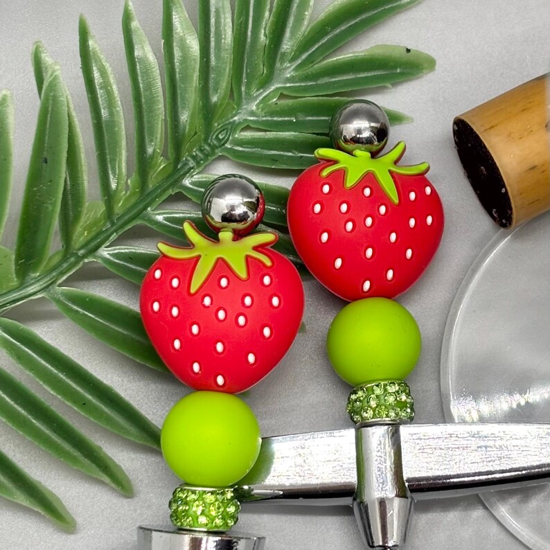 K&ouml;nnte beinhalten: Nahaufnahme von zwei Flaschenverschl&uuml;ssen im Erdbeer-Design. Jeder Verschluss hat eine rote Erdbeere mit wei&szlig;en Samen, einen gr&uuml;nen Stiel und eine silberne Kugel oben. Die Verschl&uuml;sse haben eine silberne Basis mit gr&uuml;nen und klaren Akzenten.