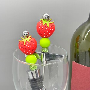 K&ouml;nnte beinhalten: Zwei Weinflaschenverschl&uuml;sse mit Erdbeermotiv. Jeder Verschluss hat eine rote Erdbeere mit wei&szlig;en Samen, eine gr&uuml;ne Perle und einen silberfarbenen Aufsatz. Die Verschl&uuml;sse werden in einem Weinglas mit einer gr&uuml;nen Weinflasche im Hintergrund gezeigt.