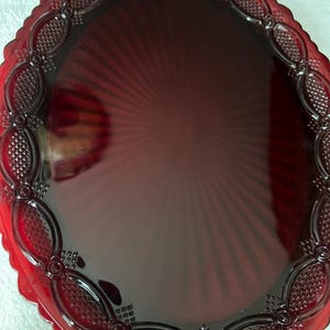 Puede incluir: Una bandeja ovalada de cristal rojo rubí con un centro liso de patrón radiante y un borde decorativo con un motivo circular y de celosía repetitivo. Los bordes de la bandeja son festoneados, creando un borde texturizado distintivo.