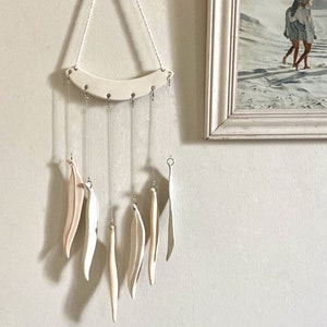 May include: A cream-colored ceramic wind chime with a curved top and dangling leaf-shaped pieces. The wind chime hangs from a white cord and is suspended in front of a light-colored wall. A framed picture is visible in the background.
