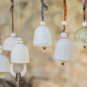 May include: A collection of white ceramic bells hanging from a branch. The bells are tied with different coloured strings and have a rustic, handmade look.