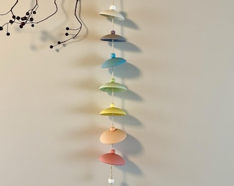 May include: A hanging mobile featuring seven pastel-colored, saucer-shaped elements strung together. Each element is a different color, including white, gray, blue, green, yellow, peach, and pink. The mobile is suspended from a thin string, with small beads between each saucer.